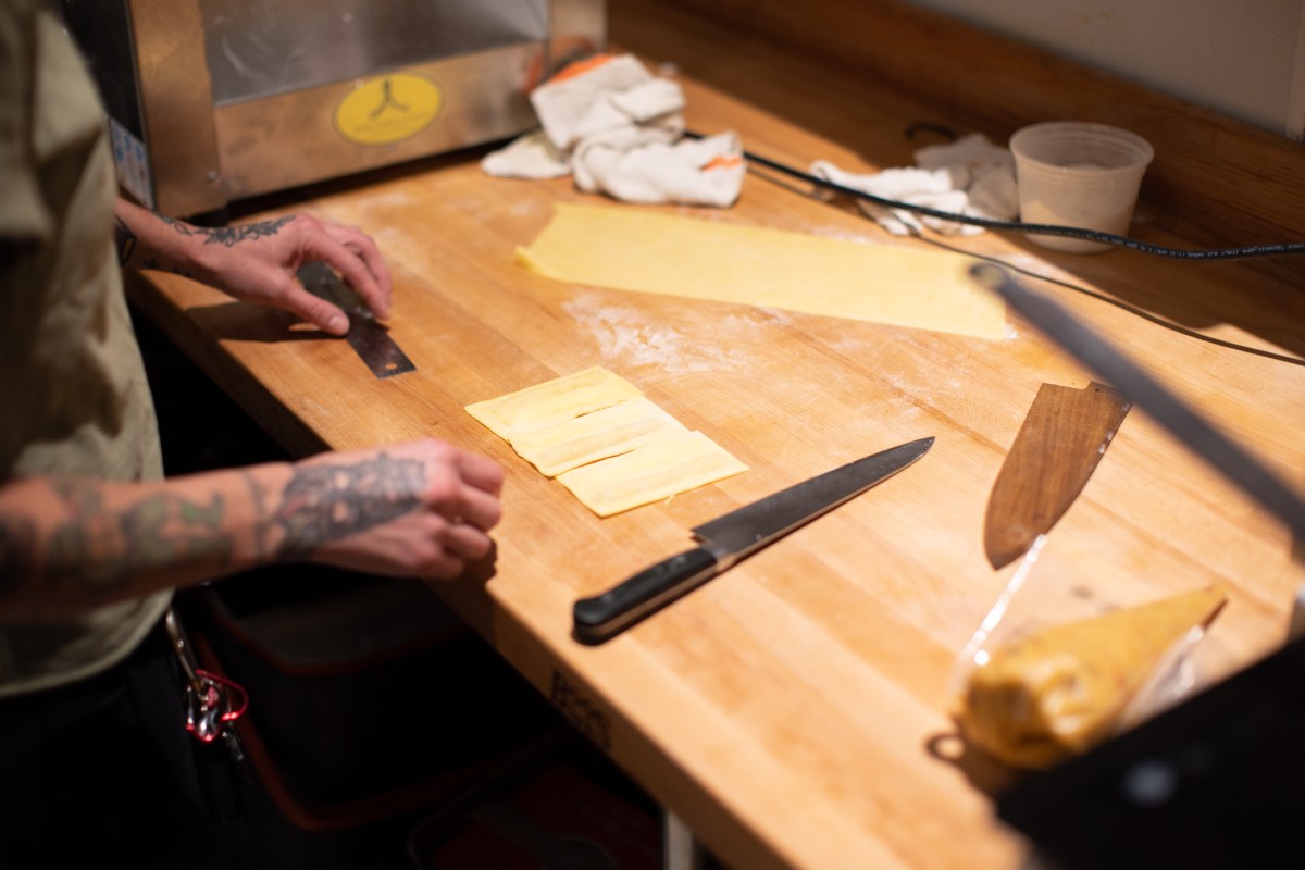 Chef's hands with tattoos cutting fresh pasta sheets on a wooden prep table