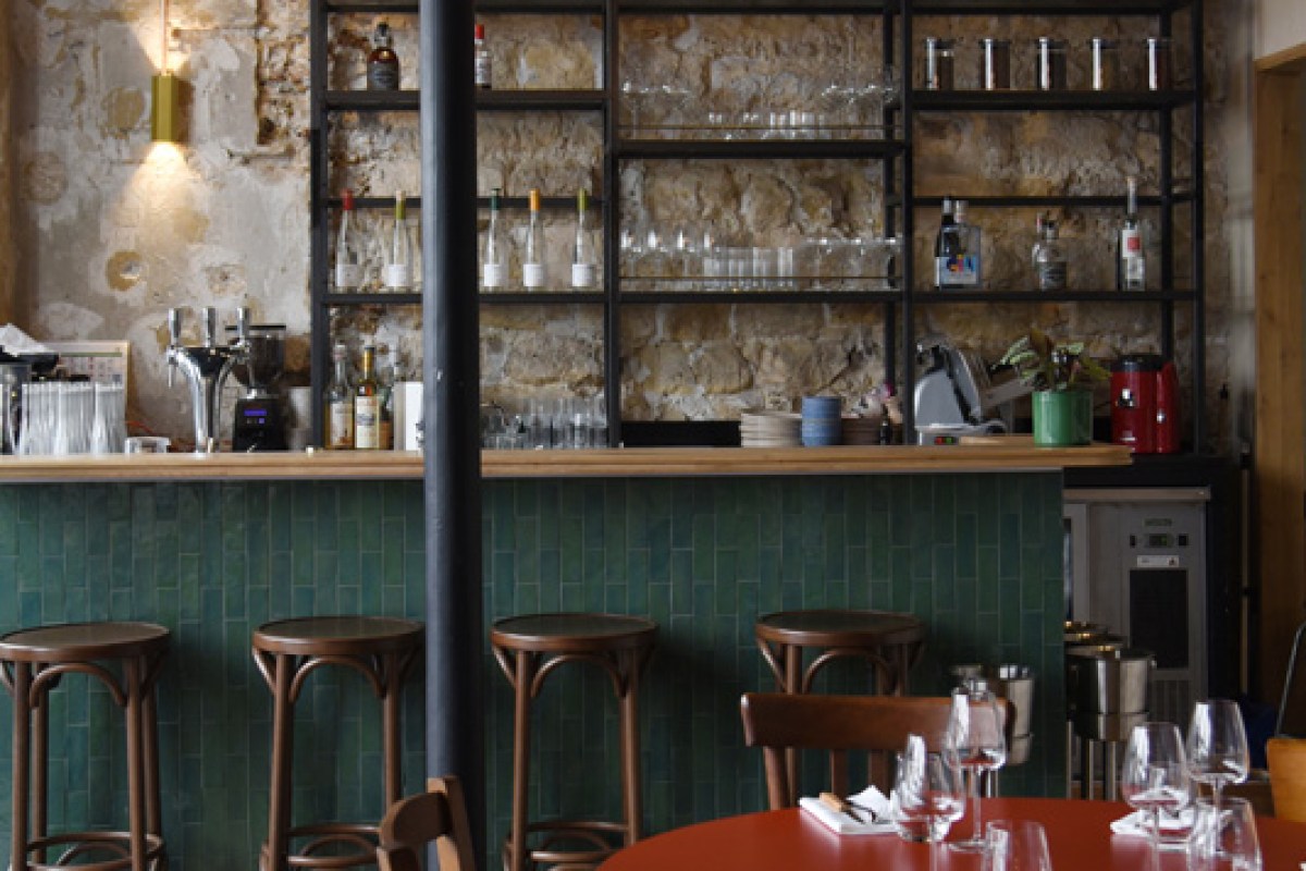 Bar area with green tiled counter, stone wall shelving with bottles and glassware, wooden bar stools, and a red table in foreground