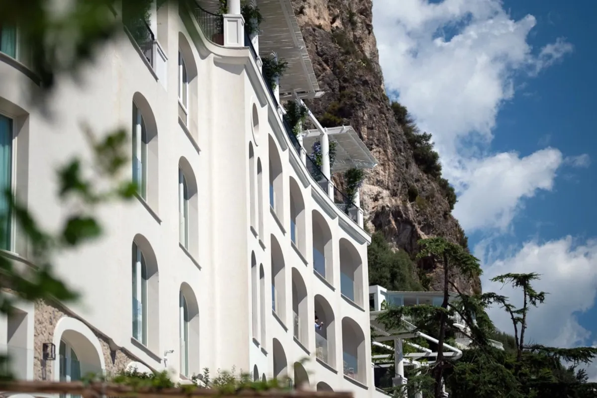White curved hotel façade with arched balconies built into a cliffside under a blue sky with clouds.