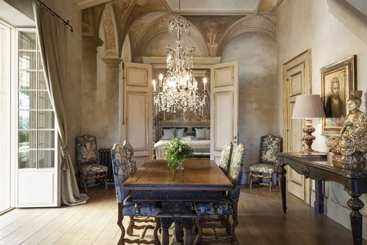 Opulent frescoed suite with chandelier, antique dining table, and glimpse of a bed through open doors.