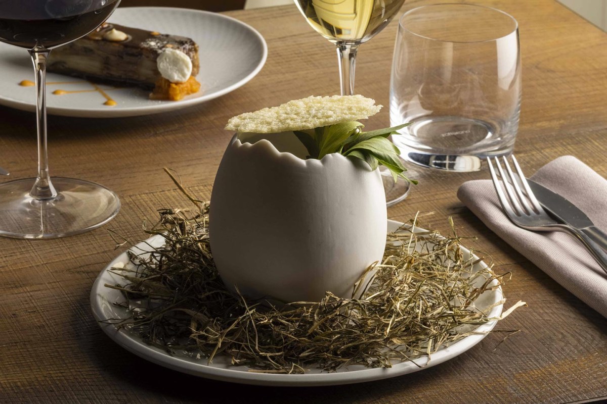 Elegant egg-shaped dish with herbs on a nest of hay, set on a wooden table with wine glasses and dessert in background