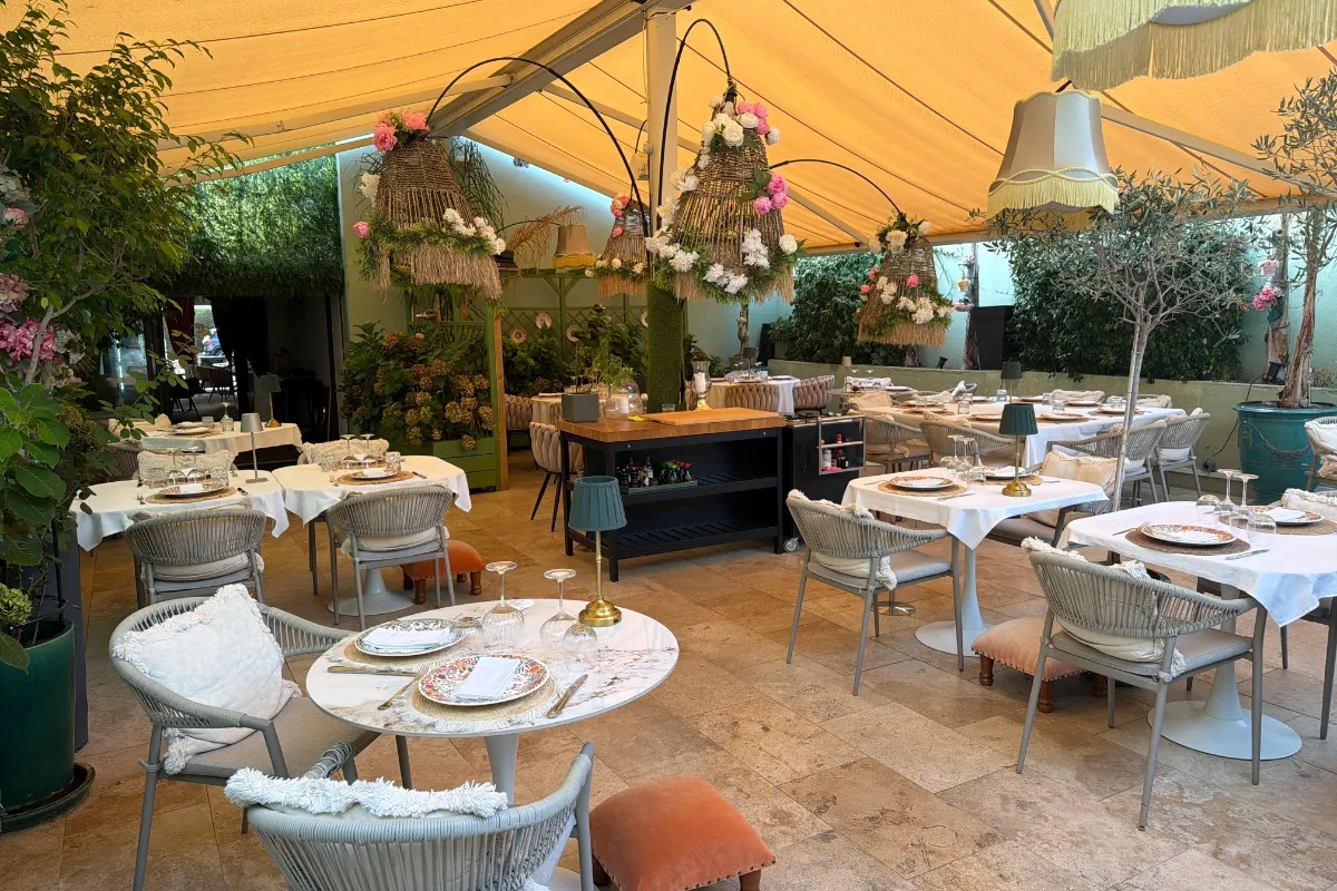 Tables laid with white cloths under the yellow awning at Brasserie Biscuit, with hanging flower baskets, olive trees and the counter behind