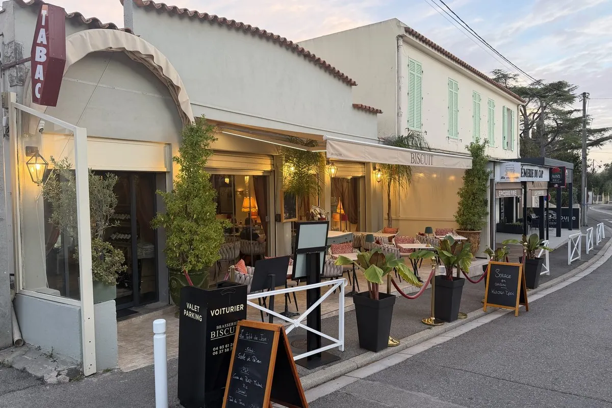 Brasserie Biscuit on Boulevard Francis Meilland at dusk, with its cream «Biscuit» awning, lit lanterns, potted palms, a terrace of laid tables, a valet parking stand and chalkboards on the pavement