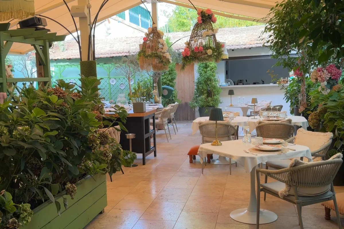 The covered garden terrace of Brasserie Biscuit on Cap d'Antibes, with laid tables, wicker chairs, planters and hanging baskets of roses and hydrangeas