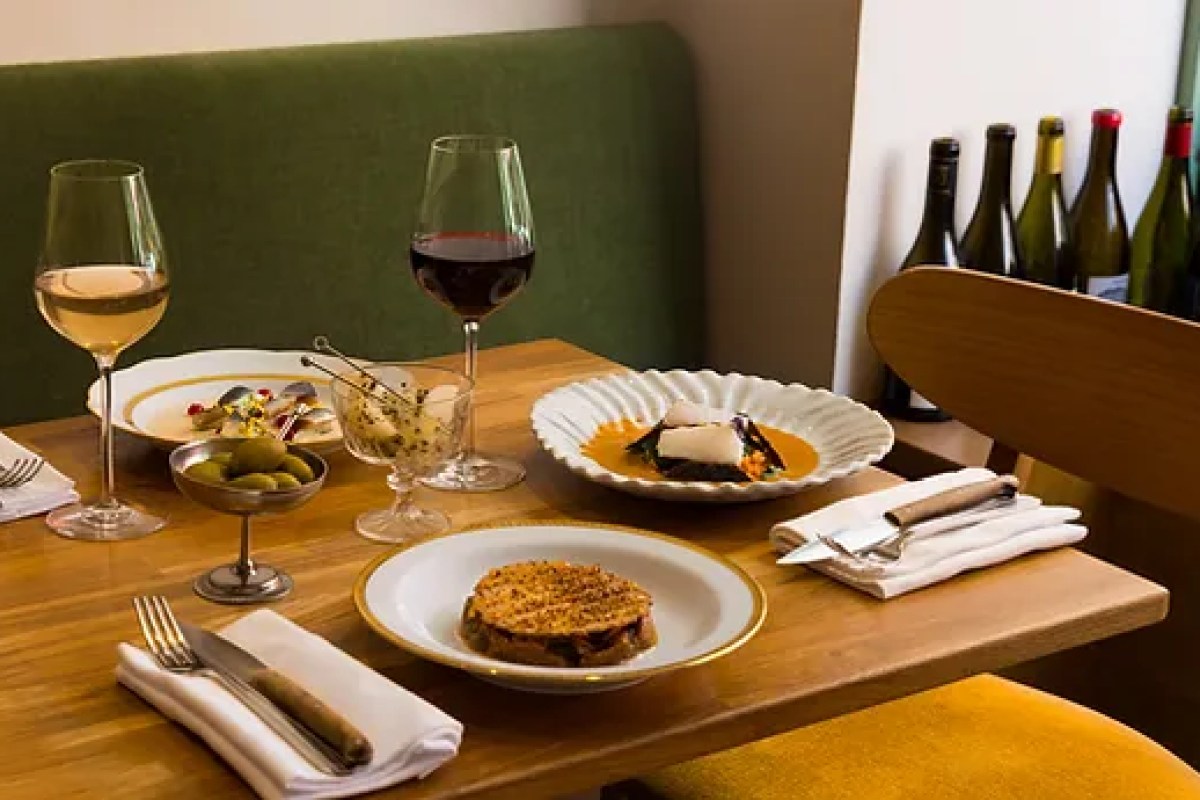 Laid wooden table with wine glasses, olives, and three plated dishes near a green banquette and wine bottles