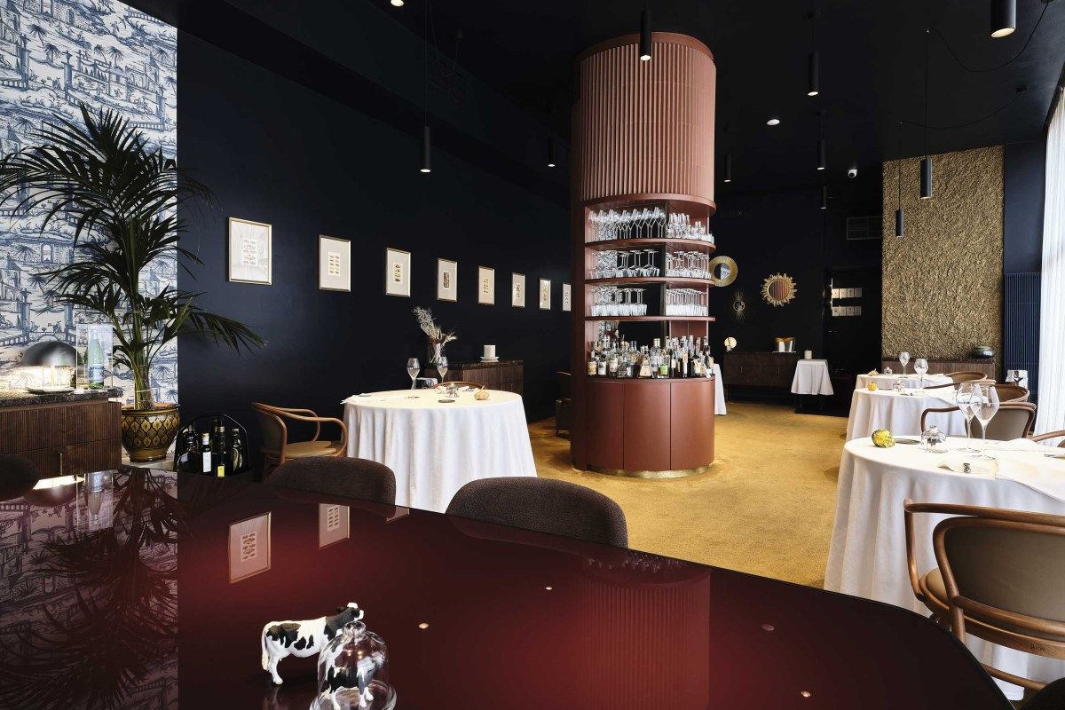 Dining room with dark walls, round white-clothed tables, a curved wood bar stocked with glassware, and framed art.
