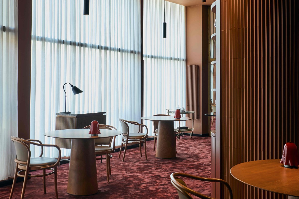 Dining area with sheer curtains, wood-paneled wall, round tables with red lamps, and burgundy carpet.