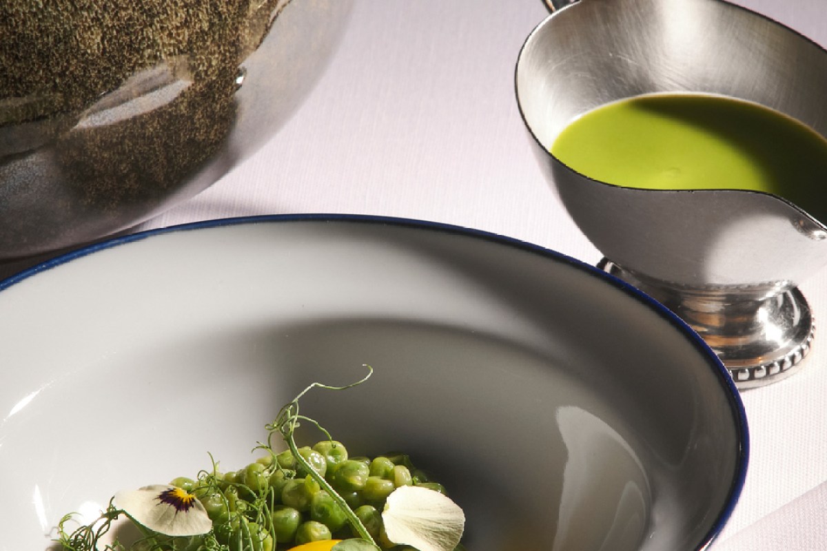 Close-up of a plated dish with peas and edible flowers, alongside a metal sauce boat of green sauce.