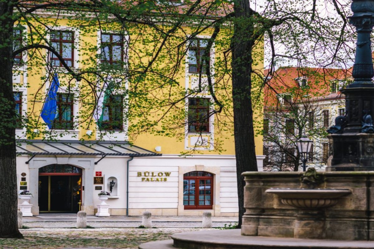 Yellow and white façade of Bülow Palais hotel with entrance canopy, seen through trees from a fountain square