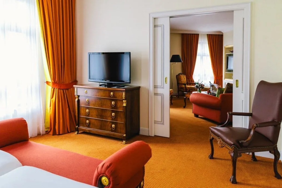 Hotel suite living area with orange sofa, wooden dresser, TV, and leather armchair, view into adjoining room