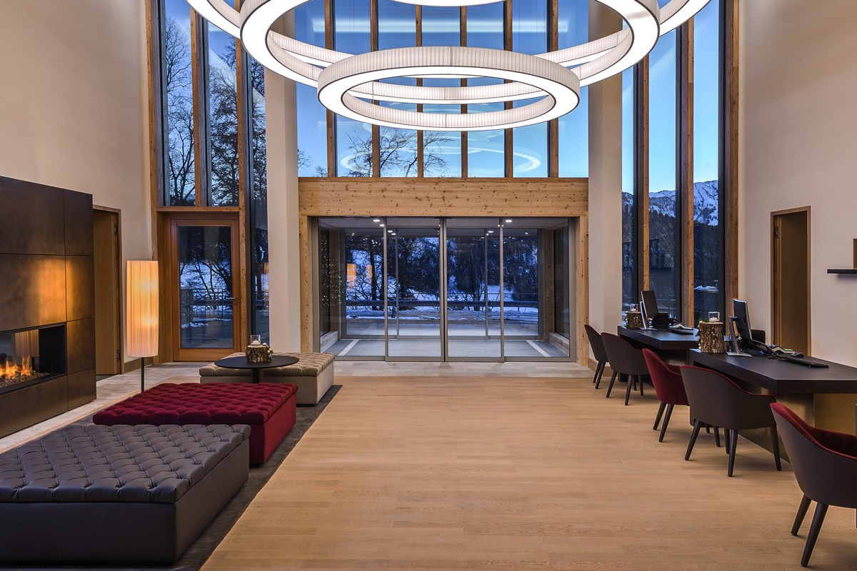 Modern hotel lobby with high wood-beamed ceiling, round pendant lights, fireplace, and reception desks