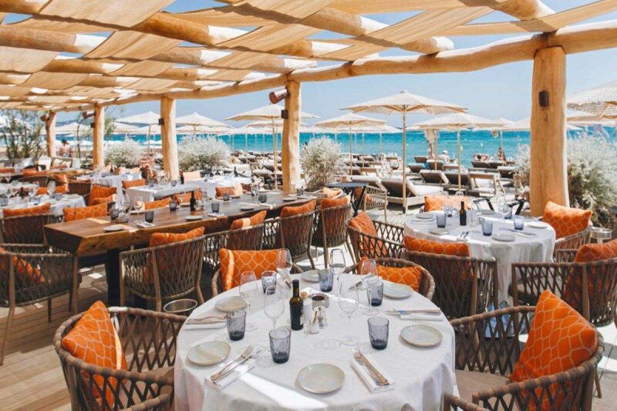Outdoor restaurant terrace with set tables, orange cushions, straw umbrellas and sea view