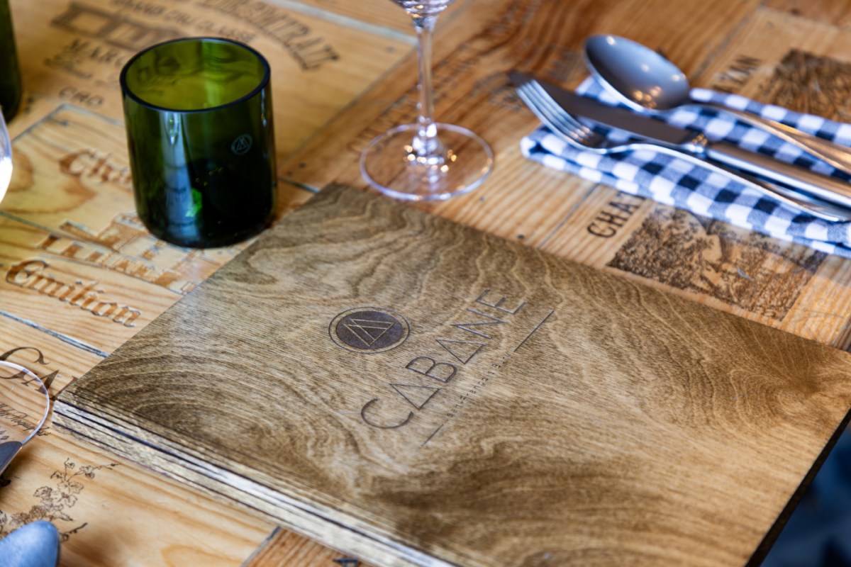 Close-up of wooden menu cover with Cabane logo, place setting, cutlery, and wine glass on patterned table