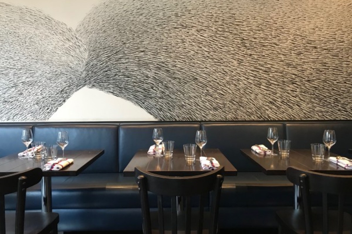 Dining room with patterned wall art above a blue banquette and dark tables set with wine glasses and napkins