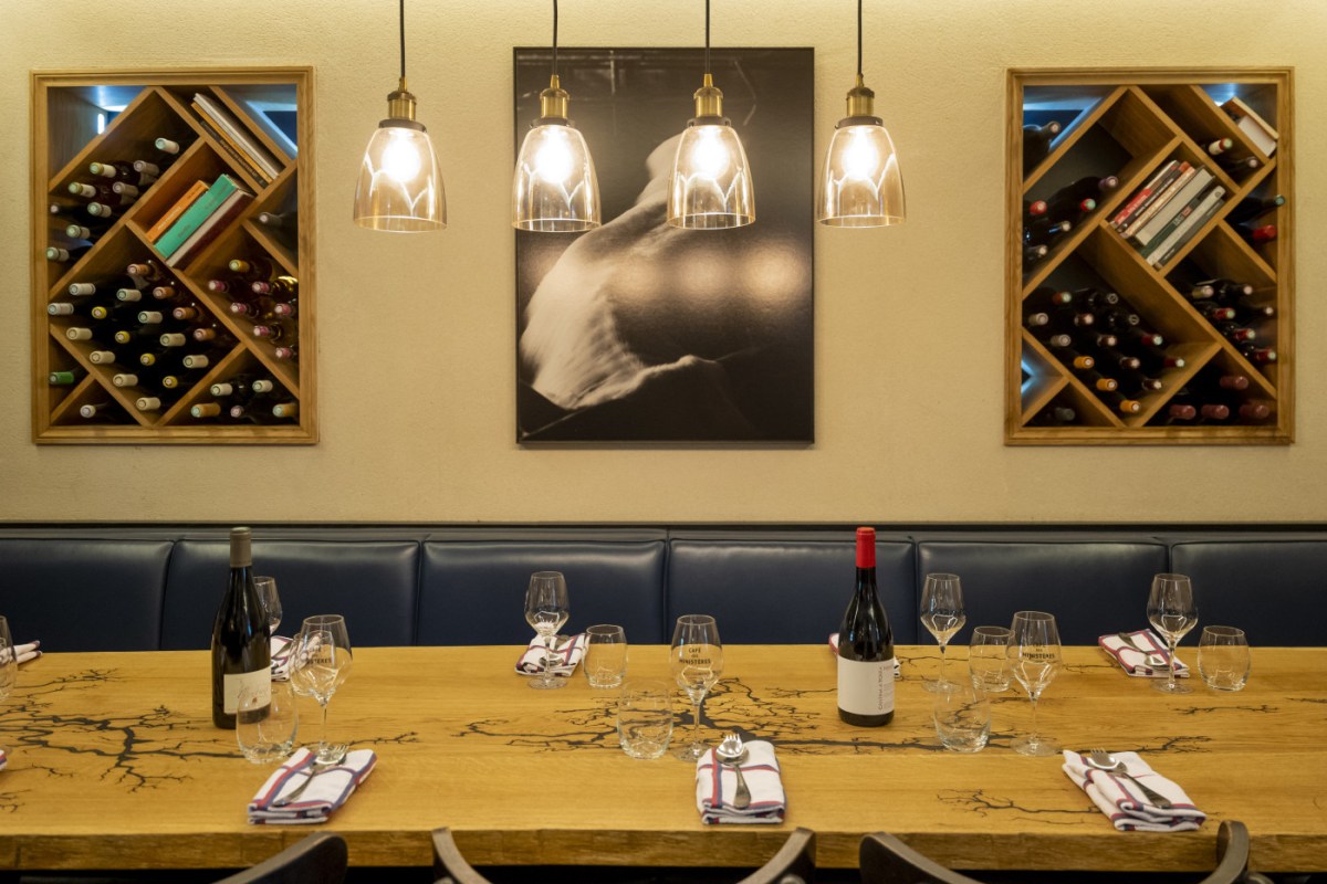 Dining room wine-rack wall with hanging lamps above a sharply laid wooden table set with wine bottles, glasses and napkins