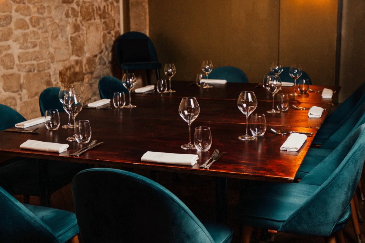 Elegant dining room table set with wine glasses, white napkins, and teal velvet chairs against a stone wall.