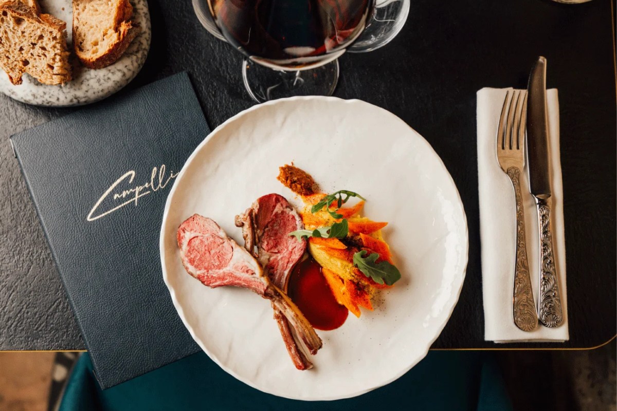 Overhead shot of lamb chops with carrots, bread, and red wine beside the Campelli menu on a dark table.