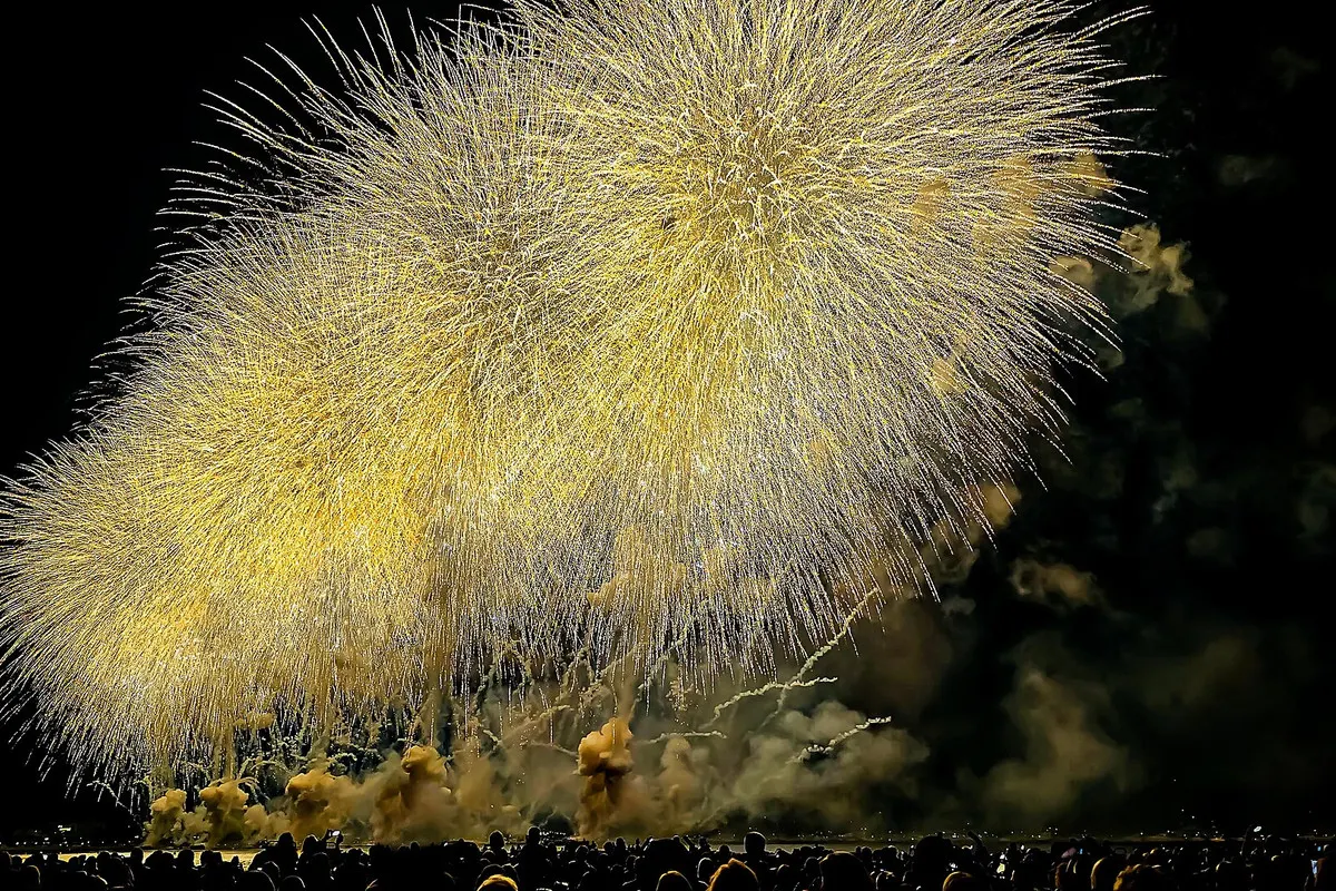Golden fireworks bursting over the bay of Cannes at night, with the silhouettes of spectators along the shore