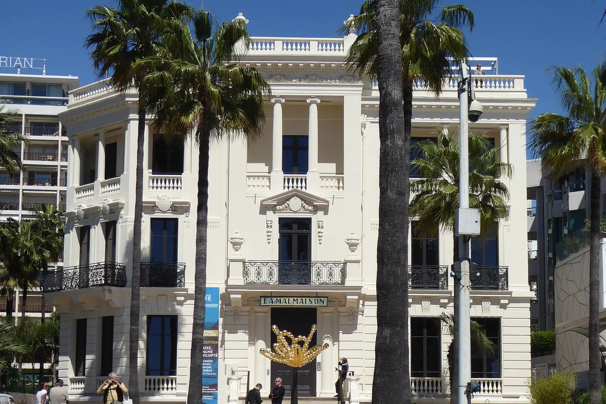 The white Belle Époque façade of La Malmaison on the Croisette in Cannes, with balustrades, wrought-iron balconies, its name above the door and palms in front