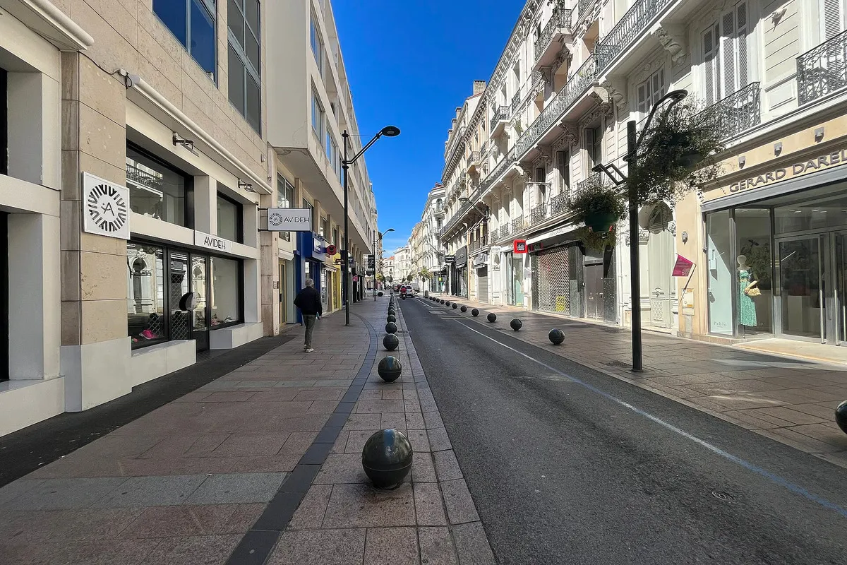 Rue d'Antibes in Cannes under a deep blue sky, a straight street of shop fronts and Belle Époque façades with wrought-iron balconies and hanging flower baskets