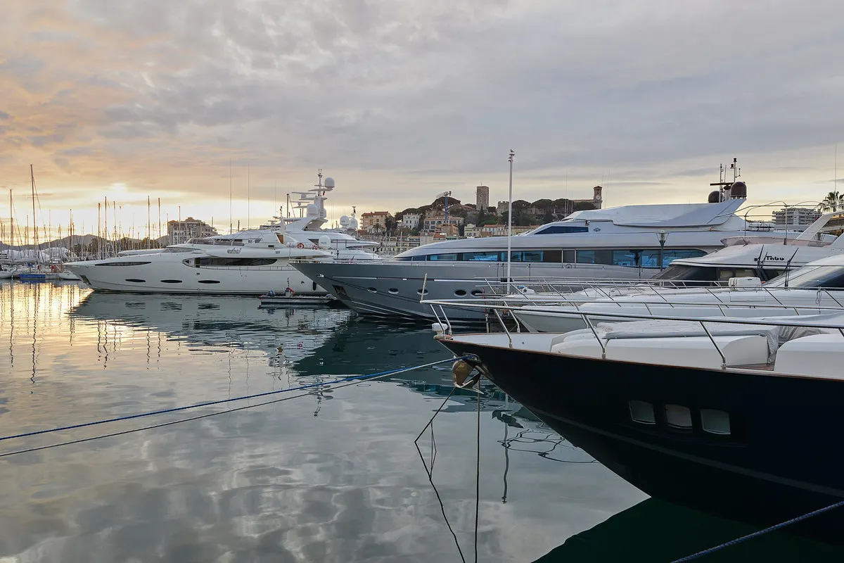 Motor yachts moored in the Vieux Port of Cannes at dawn, reflected in still water, with Le Suquet and its tower on the hill behind