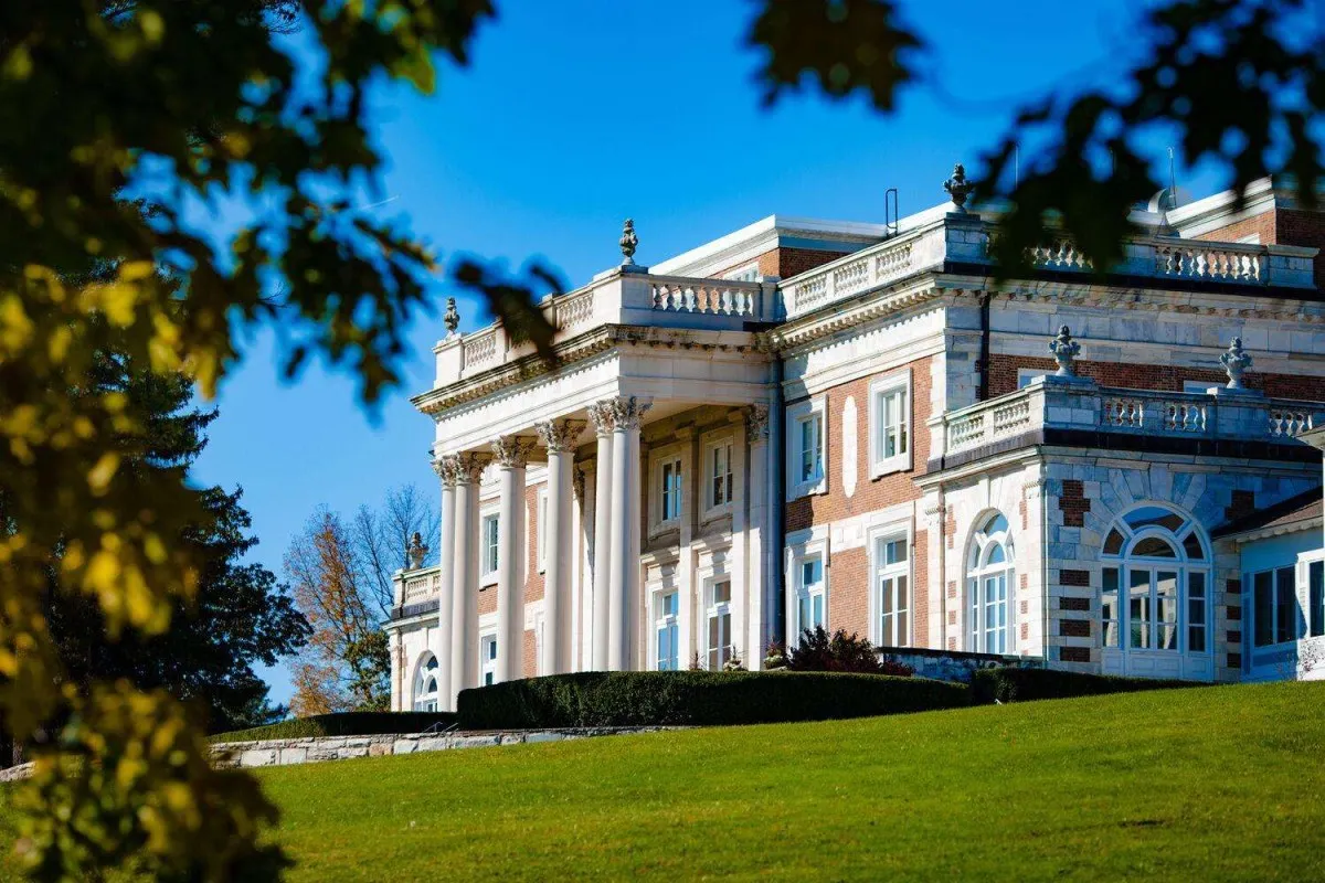 Grand brick and white stone mansion facade with columns, framed by autumn leaves under blue sky, green lawn in front