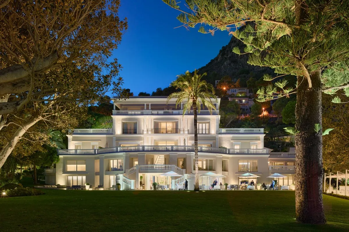 The white Manor House at Cap Estel in Èze-Bord-de-Mer lit at dusk behind a lawn and pine trees; La Table du Cap Estel is on its ground floor