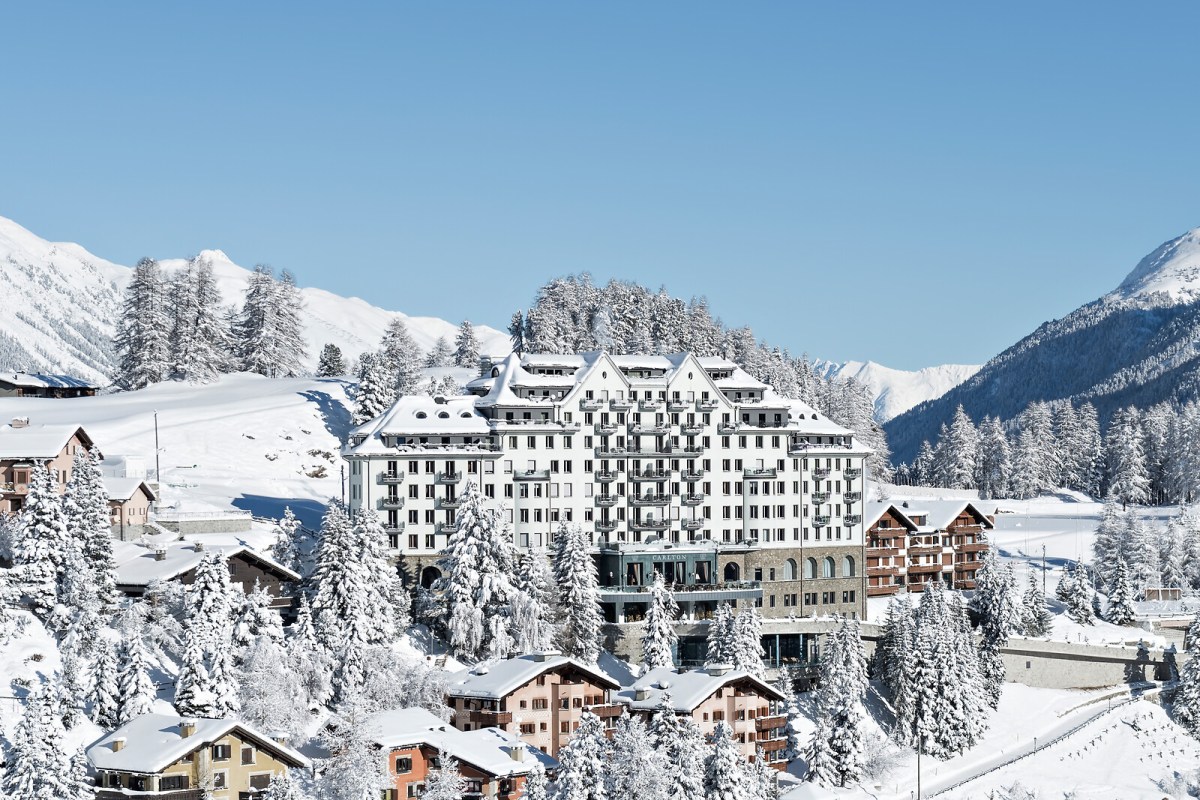 Snow-covered Carlton Hotel St. Moritz facade with visible signage, surrounded by pine trees and mountains under blue sky