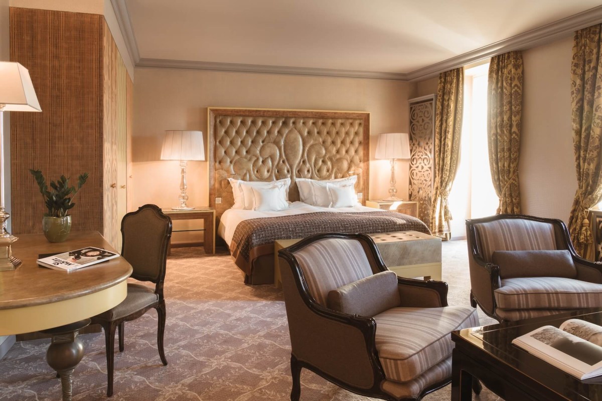 Elegant hotel bedroom with tufted gold headboard, striped armchairs and patterned carpet