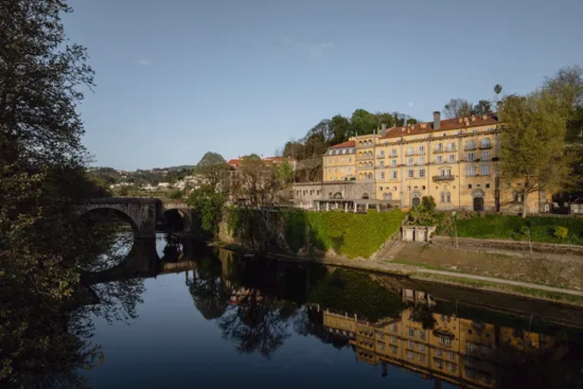 Casa da Calçada hotel building seen across a river with a stone bridge and reflections, surrounded by trees