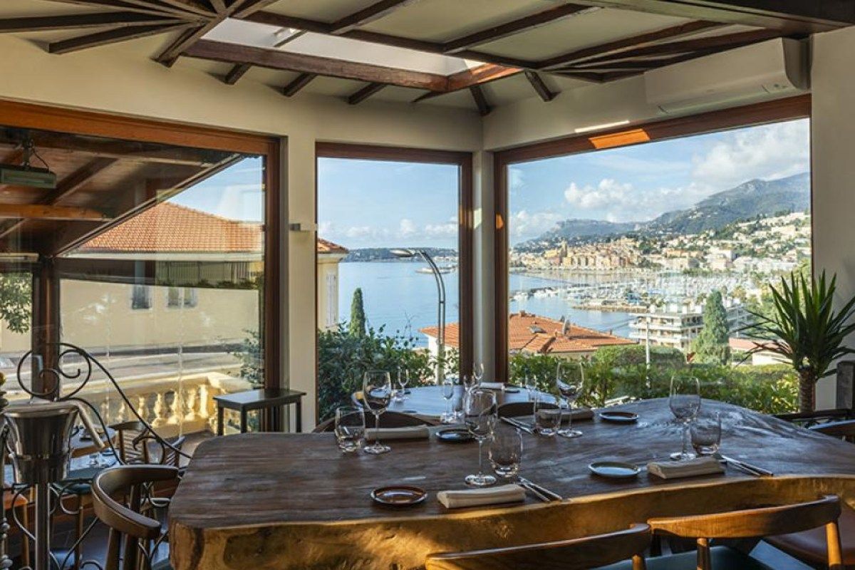 Dining room with wooden table set for a meal, large windows overlooking Menton's harbor and old town