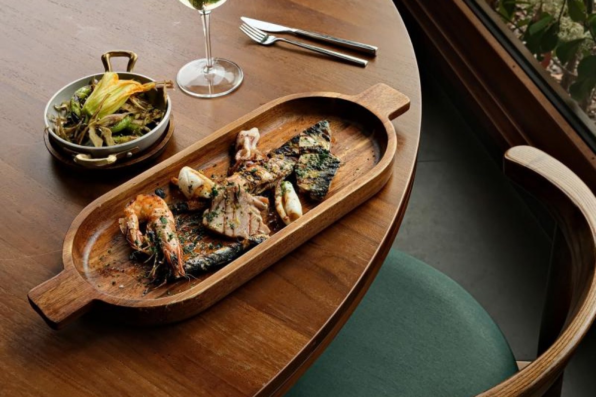 Wooden board with grilled shrimp, fish, and calamari on a restaurant table with a glass of white wine