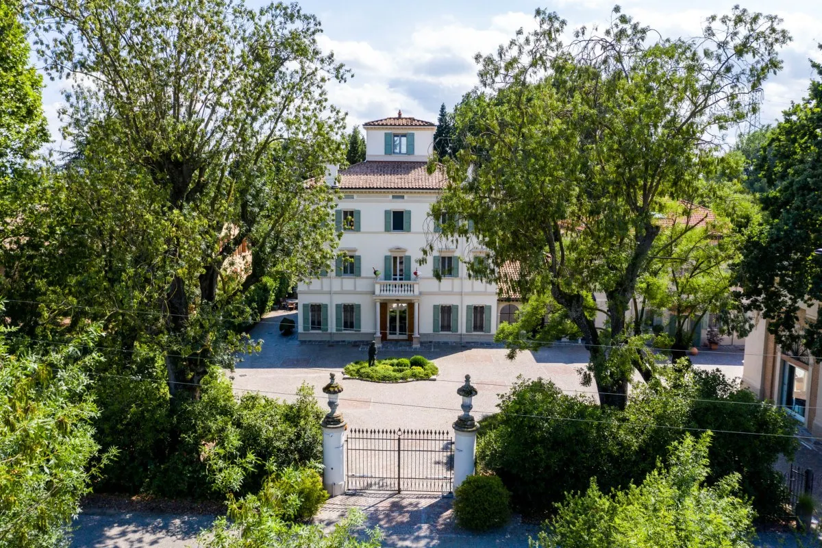Aerial view of Casa Maria Luigia's cream façade with green shutters, framed by trees and a gated entrance drive.
