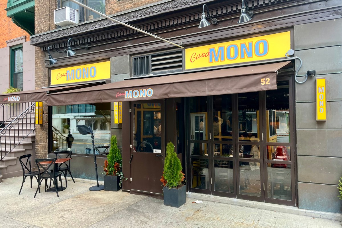 Casa Mono restaurant storefront with yellow signs, brown awning, entrance door, and small sidewalk table