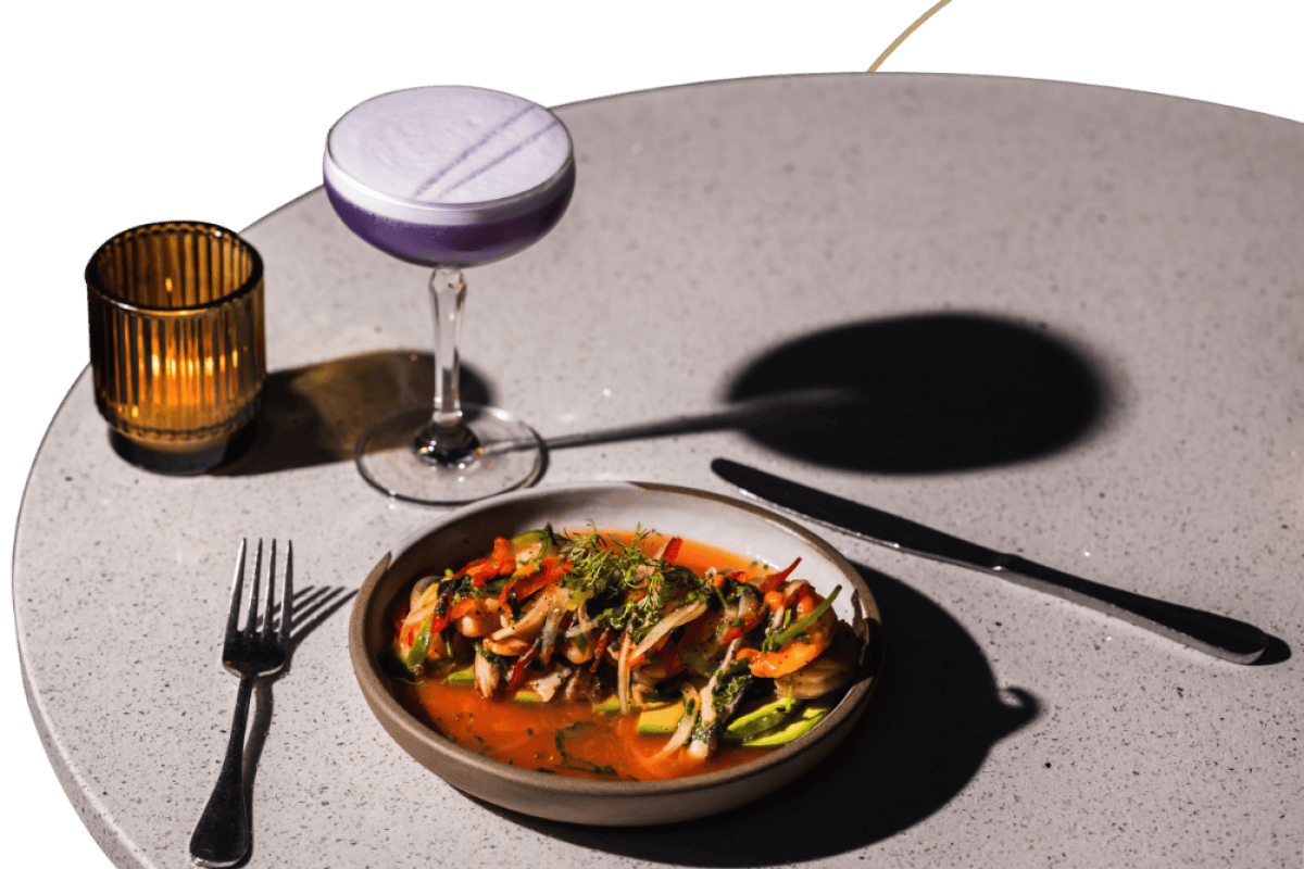 Plated seafood dish with herbs beside a purple cocktail and candle on a round speckled table