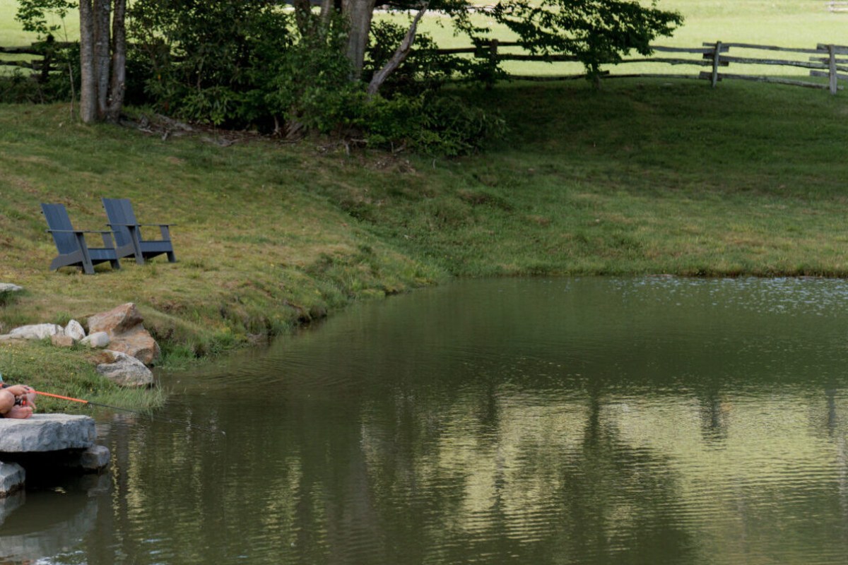 A calm pond with two blue Adirondack chairs on the grassy bank, bordered by a wooden split-rail fence at the ranch