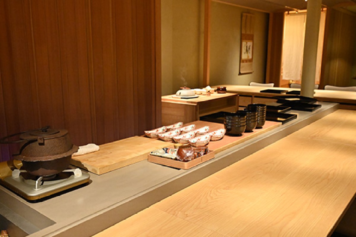Interior counter and dining area of a Japanese restaurant with wooden counter, iron kettle, lacquer bowls, and hanging scroll