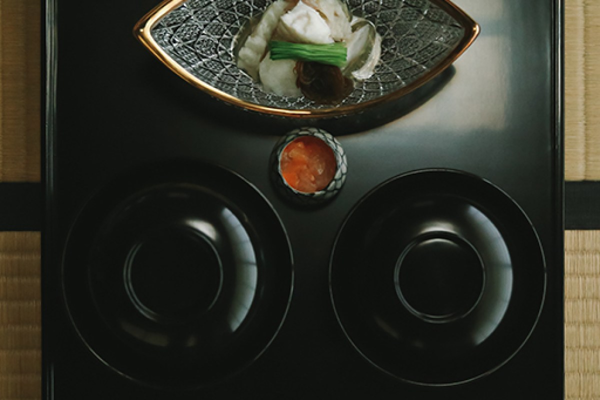 Overhead shot of a black lacquered tray with a shell-shaped dish holding delicate seafood, garnish, and small sauce cup on tatami mat