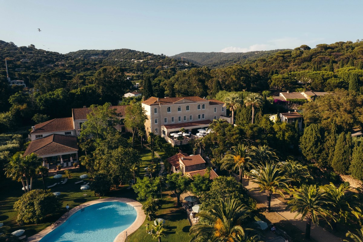 Aerial view of the hotel building surrounded by palm trees, gardens and a pool, with hills in the background