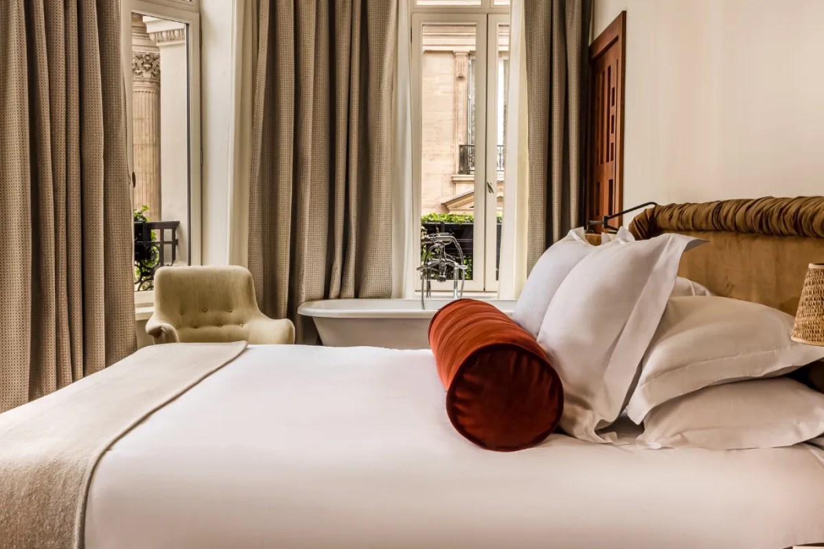 Guest room with bed, red bolster pillow, armchair, and freestanding bathtub by window overlooking Paris street