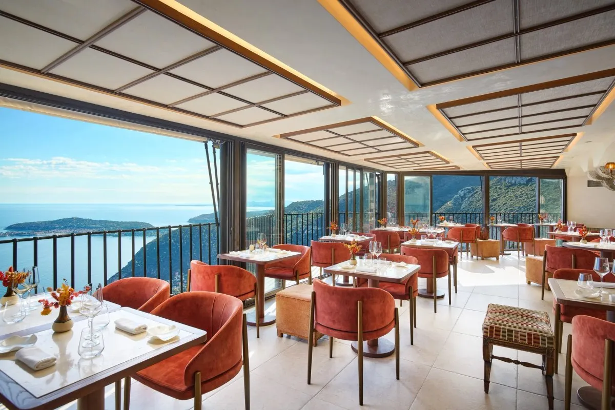 The dining room of La Table Gastronomique at Château Eza, with coral armchairs and floor-to-ceiling windows overlooking the Mediterranean and Cap Ferrat