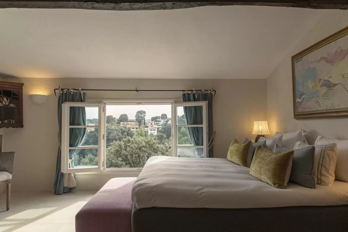 Elegant guest room with large bed, soft pillows, and a window overlooking green hillside and villas
