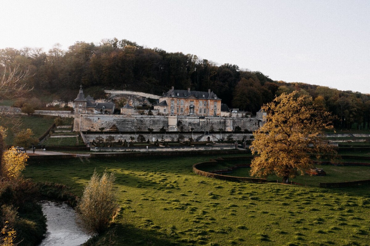 Wide view of Château Neercanne nestled against a wooded hillside with gardens and a stream in the foreground
