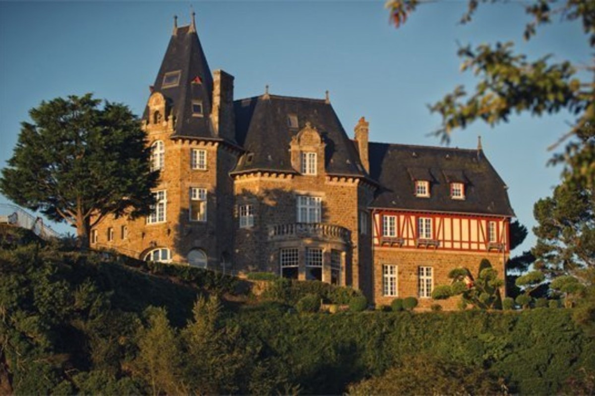 Full view of the château's stone façade with turrets, half-timbered wing, and chimneys at golden hour, surrounded by hedges and trees