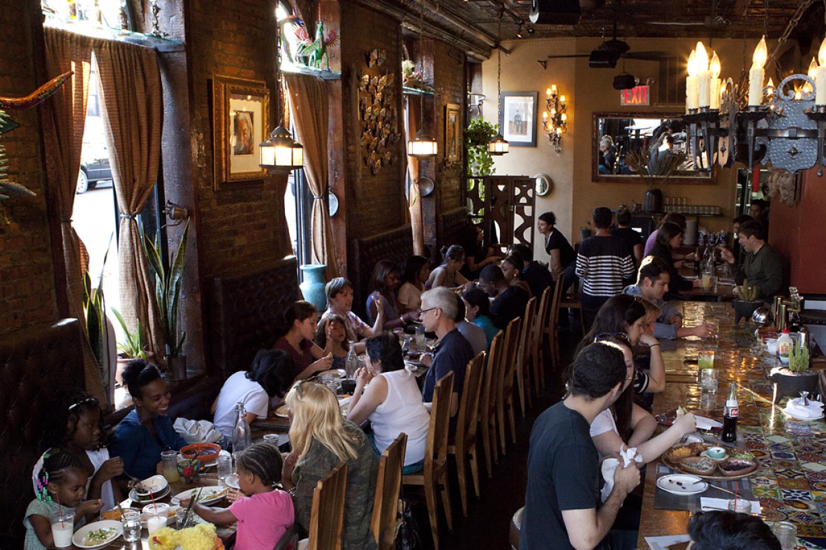 Wide view of Chavela's rustic dining room with brick walls, hanging lights and bar, diners seated