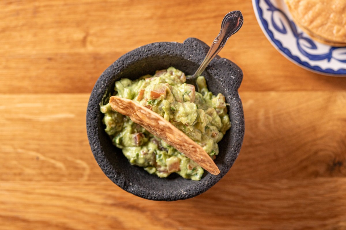 Molcajete bowl of fresh guacamole with tortilla chip and spoon, blue-and-white plate of chips beside it