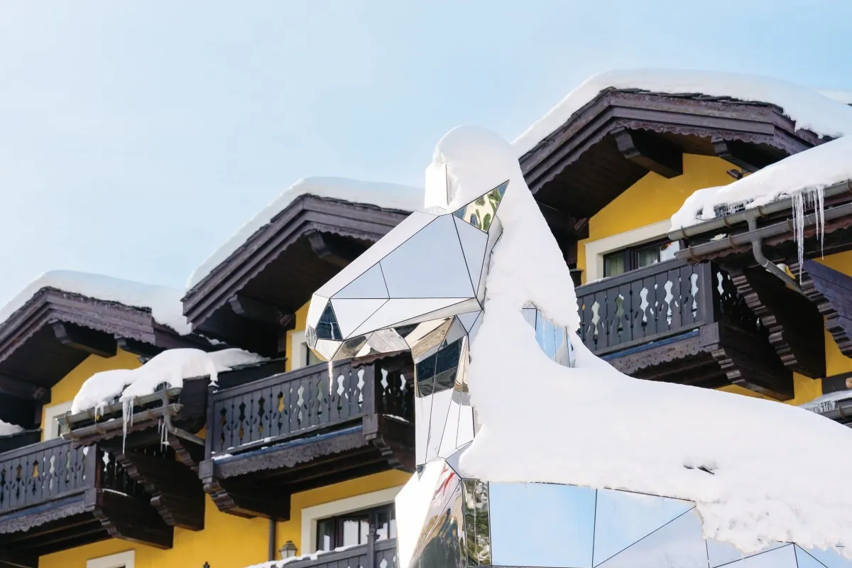 Snow-covered chalet-style hotel facade with wooden balconies and a mirrored horse head sculpture in front