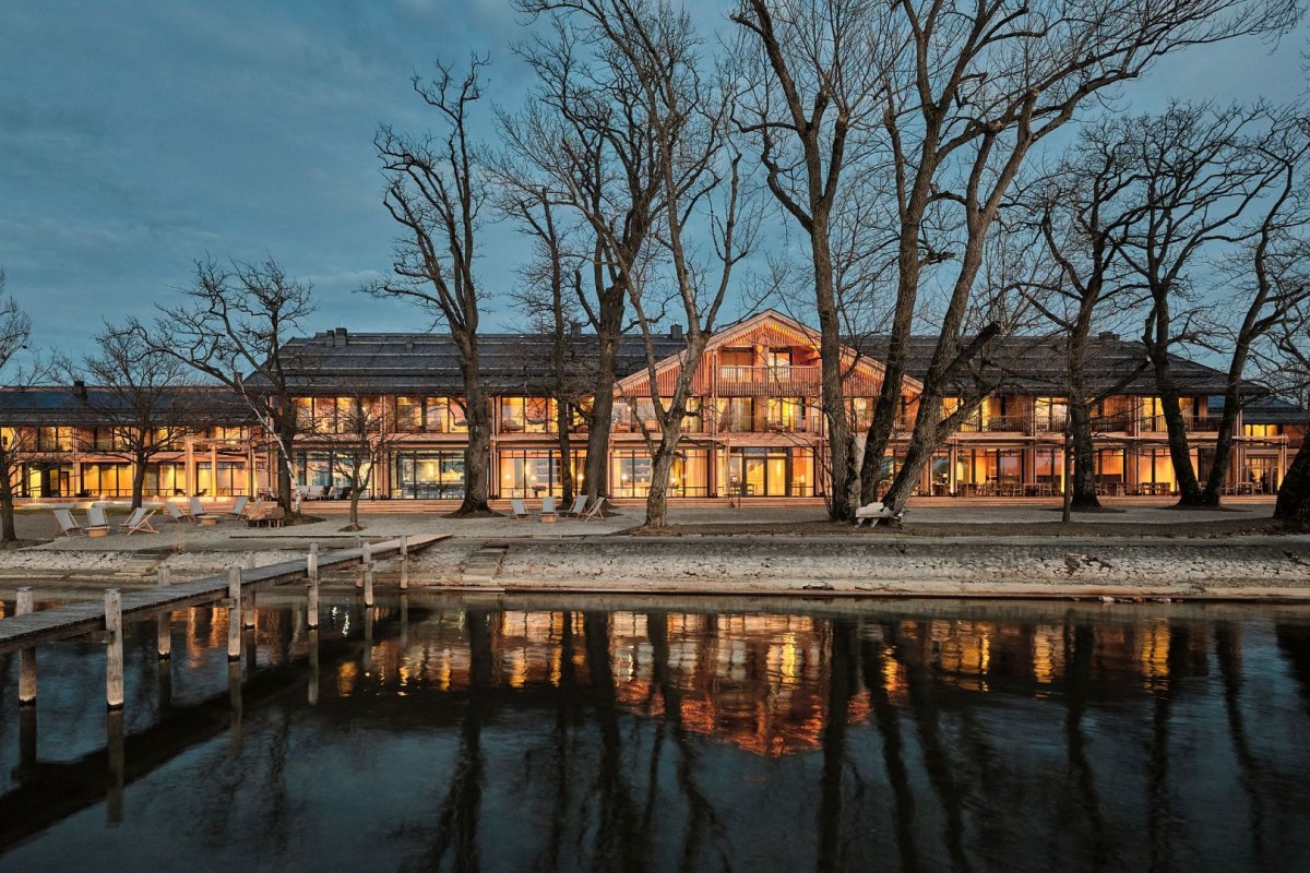 Lakeside hotel building illuminated at dusk, wooden façade with balconies, surrounded by bare trees, reflected in calm water with a dock in foreground.
