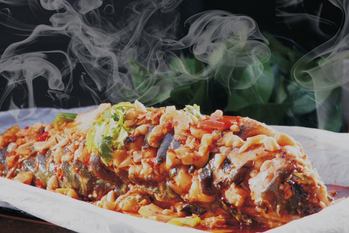 Whole grilled fish topped with chili, garlic, and herbs, plated with visible steam.