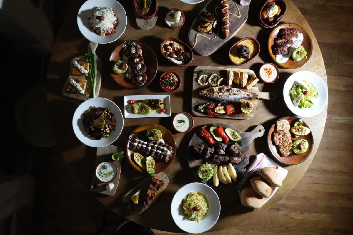 Overhead view of a round wooden table filled with assorted Balkan dishes, grilled meats, fish, and salads
